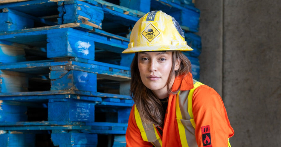 A woman wearing a hard hat, work boots, and orange high-visibility coveralls while sitting on a stack of pallets.