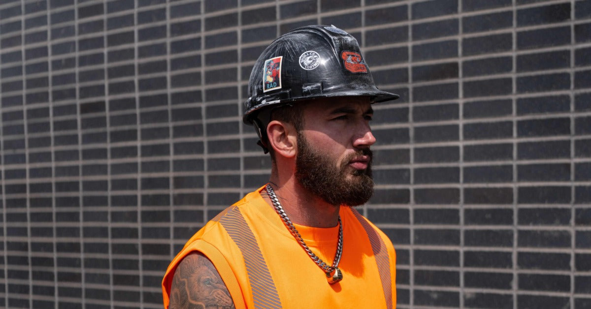 A close-up of a man wearing a black hard hat and a sleeveless orange high-visibility shirt with reflective stripes.