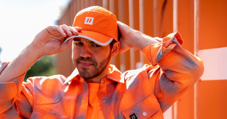 A person wearing an orange hi-vis hat, shirt, and flannel shirt while walking next to an orange building outside.