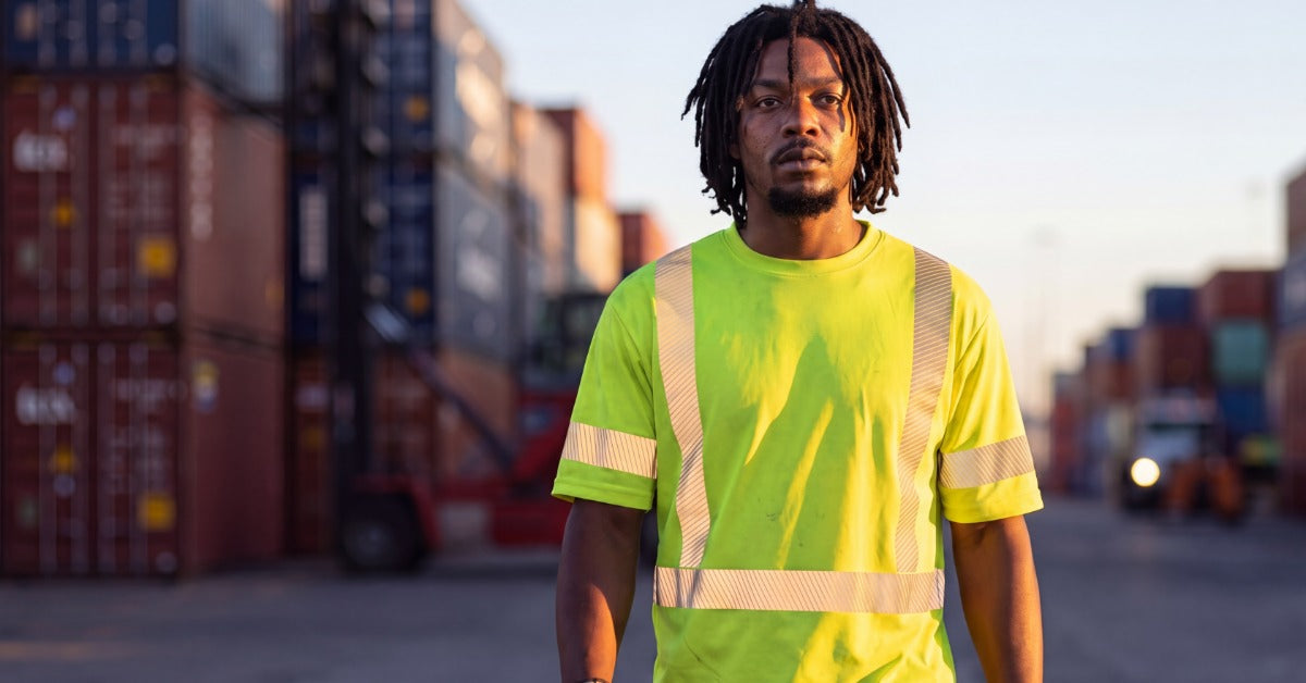 A worker wearing a high-visibility shirt and safety gloves while walking through a shipping container yard.