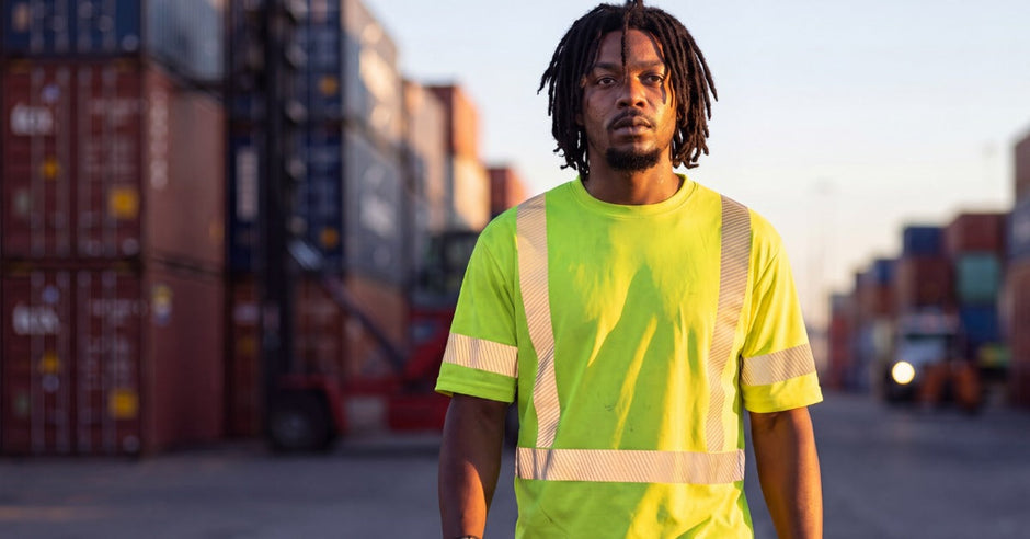 A worker wearing a high-visibility shirt and safety gloves while walking through a shipping container yard.