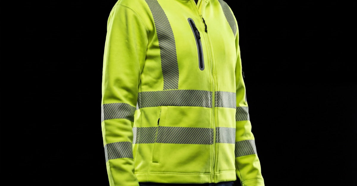 A woman wearing a high-visibility reflective hoodie and dark work pants while standing against a black background.