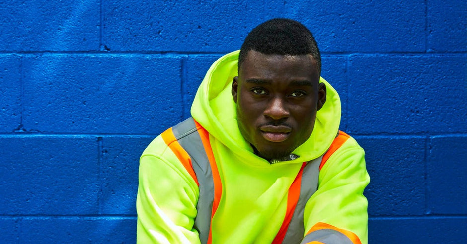 A man in a hi-vis yellow reflective hoodie and jeans sitting against a bright blue brick wall while looking at the camera.
