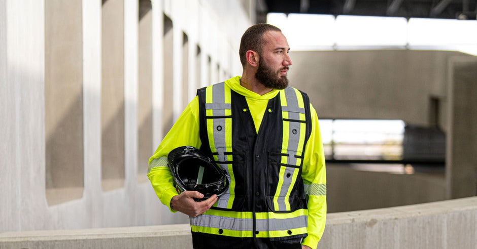 A construction worker wearing a high-visibility safety vest and work clothes while holding a black hard hat.