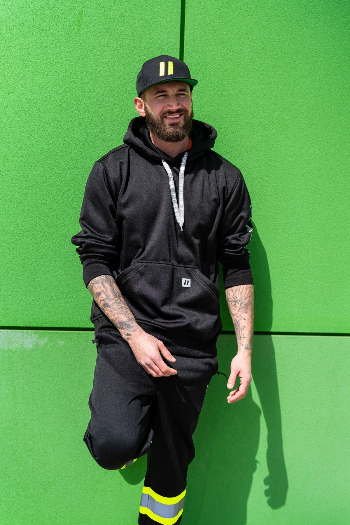 A smiling bearded man in a black hoodie, black work pants, and a cap leaning against a bright green wall.