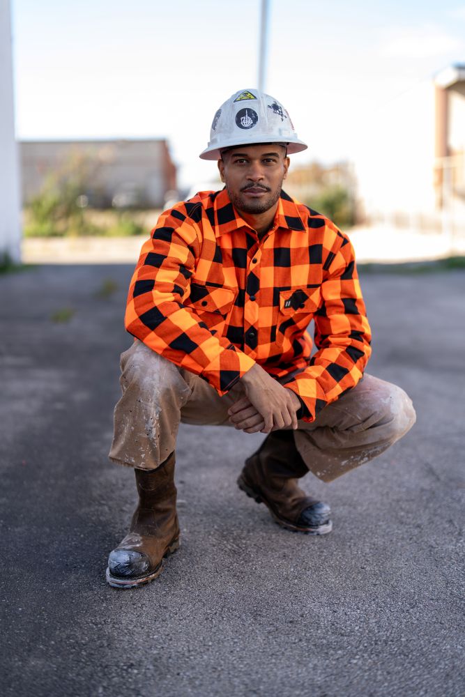 A construction worker in a white hard hat, orange and black plaid shirt, work pants, and boots crouching outdoors.