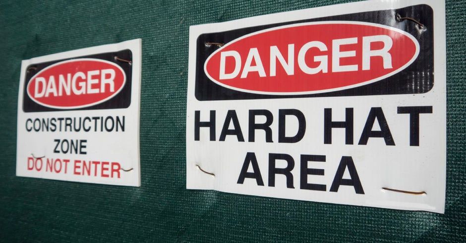 A green construction fence with two signs saying "danger, construction zone" and "danger, hard hat area."