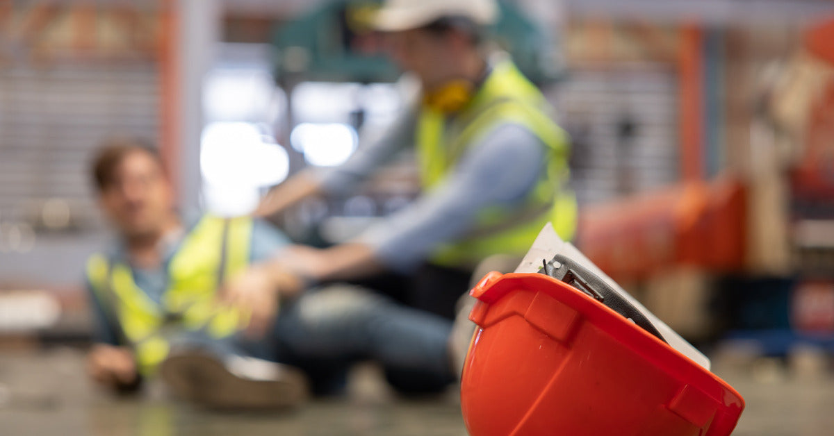 Close-up of a red hard hat with an injured factory worker and someone tending to them in the background.